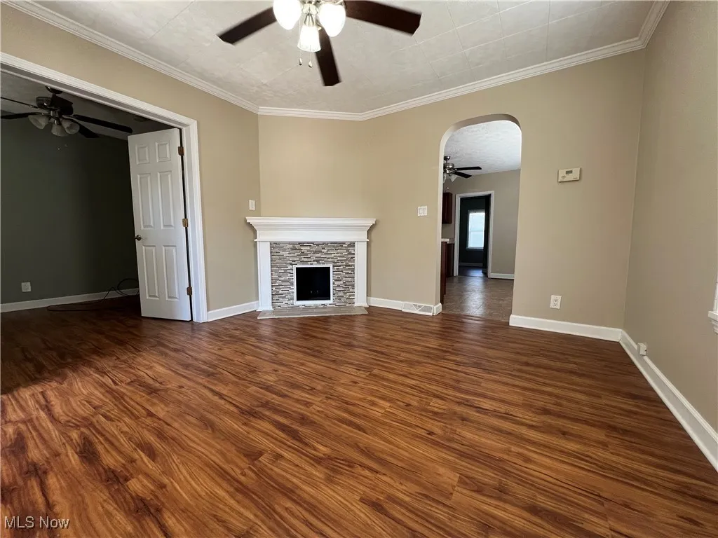 Unfurnished living room featuring a ceiling fan, dark wood finished floors, a fireplace, arched walkways, and ornamental molding