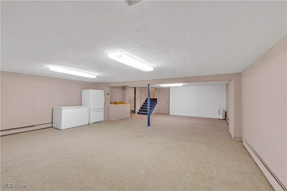 Finished below grade area with baseboard heating, a textured ceiling, light colored carpet, freestanding refrigerator, and stairway