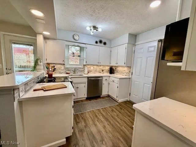 Kitchen featuring white cabinetry, a textured ceiling, stainless steel dishwasher, dark LVP dark walnut wood-style floors, and a quartz peninsula.