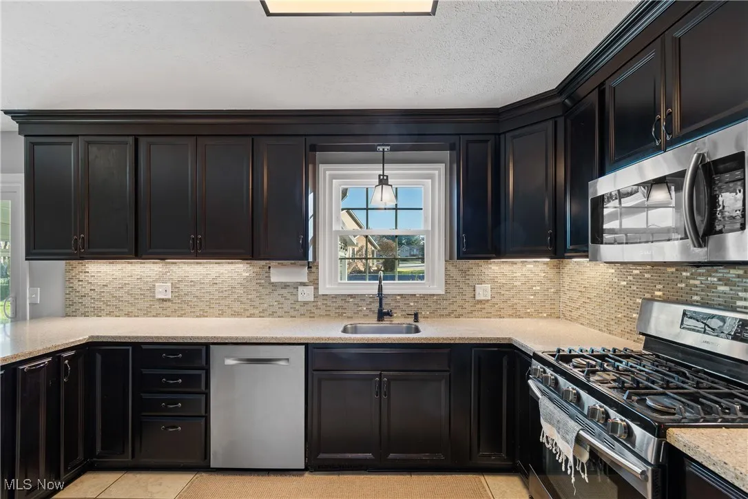 Kitchen with appliances with stainless steel finishes, decorative backsplash, light stone countertops, dark cabinetry, and a textured ceiling