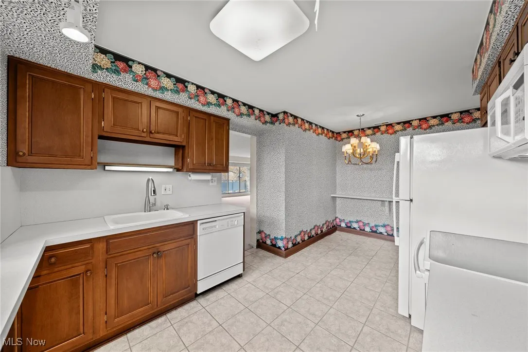 Kitchen featuring white appliances, light countertops, brown cabinetry, pendant lighting, and wallpapered walls