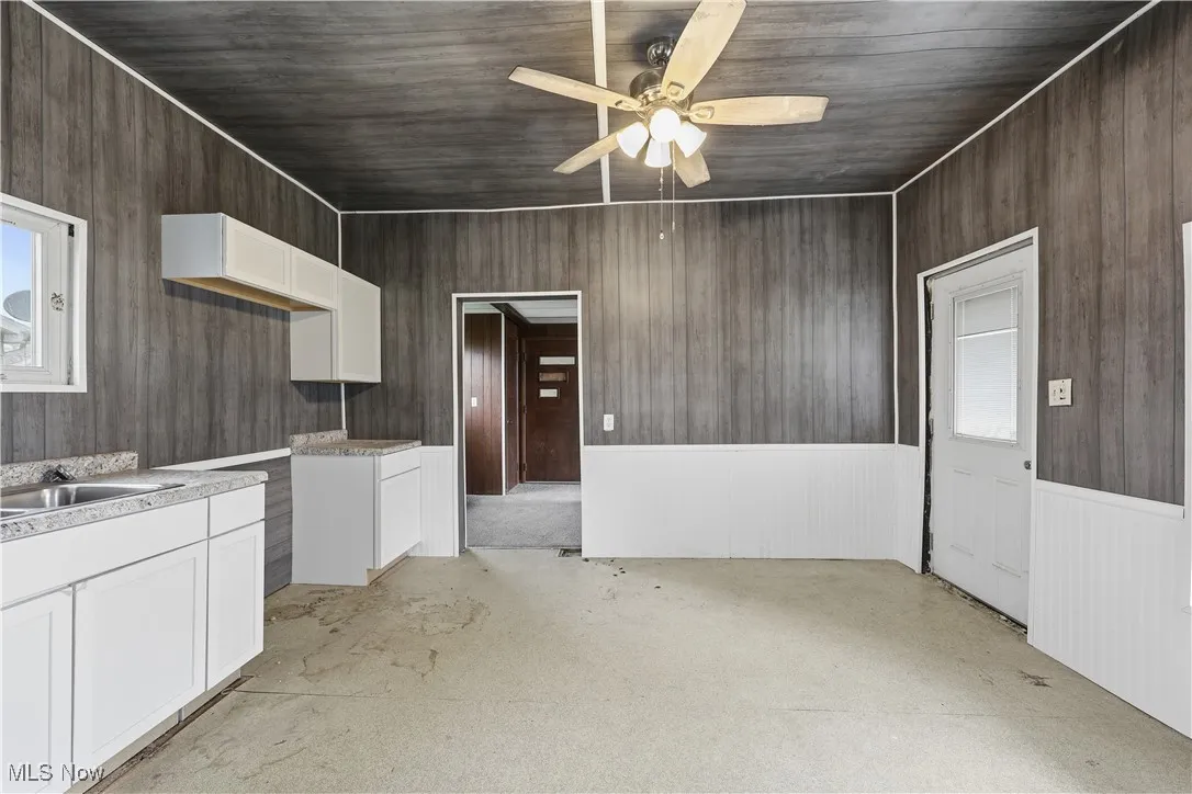 Kitchen featuring white cabinetry, wooden walls, light countertops, a ceiling fan, and wainscoting