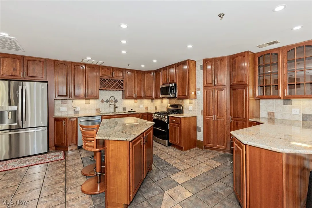 Kitchen with glass insert cabinets, brown cabinetry, appliances with stainless steel finishes, light stone counters, and recessed lighting
