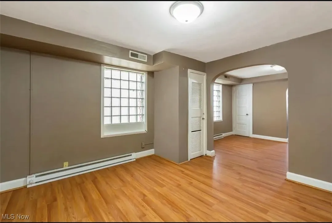 Empty room with a baseboard radiator, arched walkways, light wood-type flooring, and healthy amount of natural light