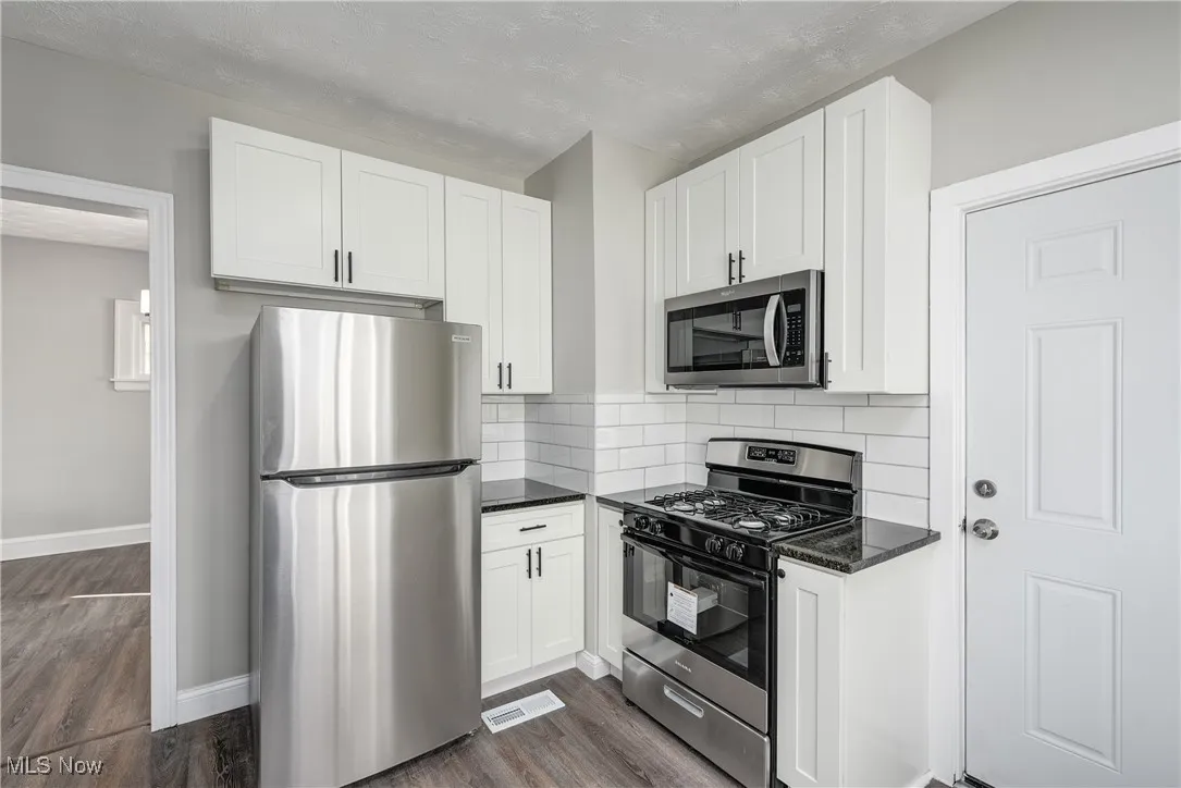 Kitchen with stainless steel appliances, white cabinets, dark stone counters, decorative backsplash, and dark wood-style flooring
