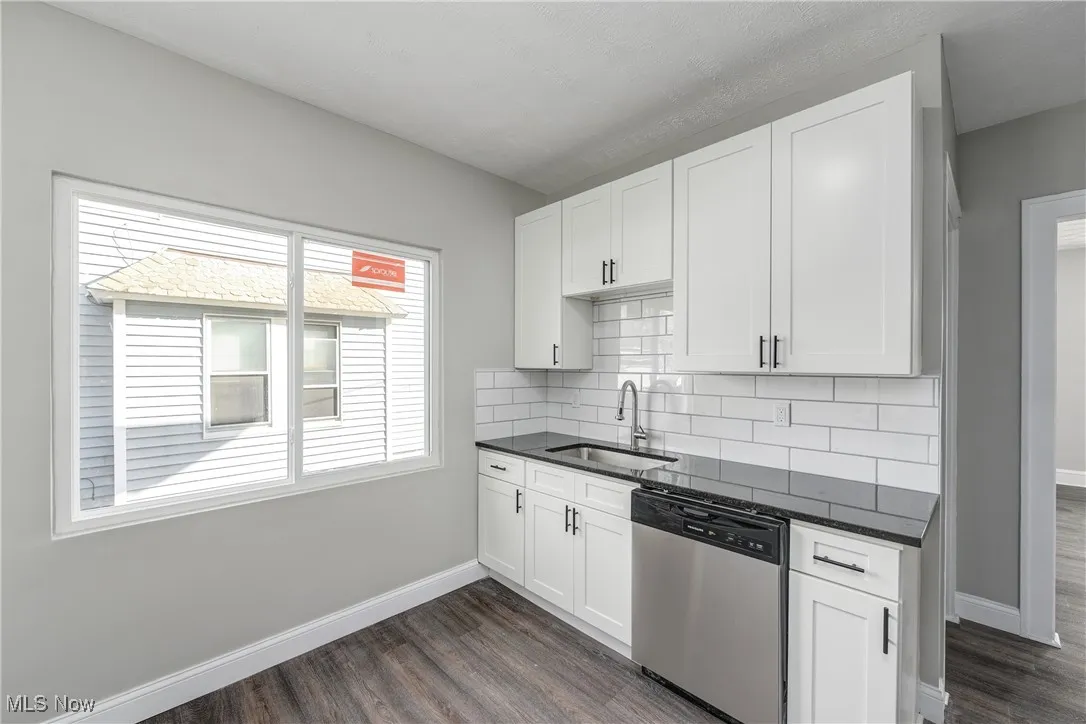 Kitchen with stainless steel dishwasher, white cabinetry, dark stone counters, backsplash, and dark wood-style floors