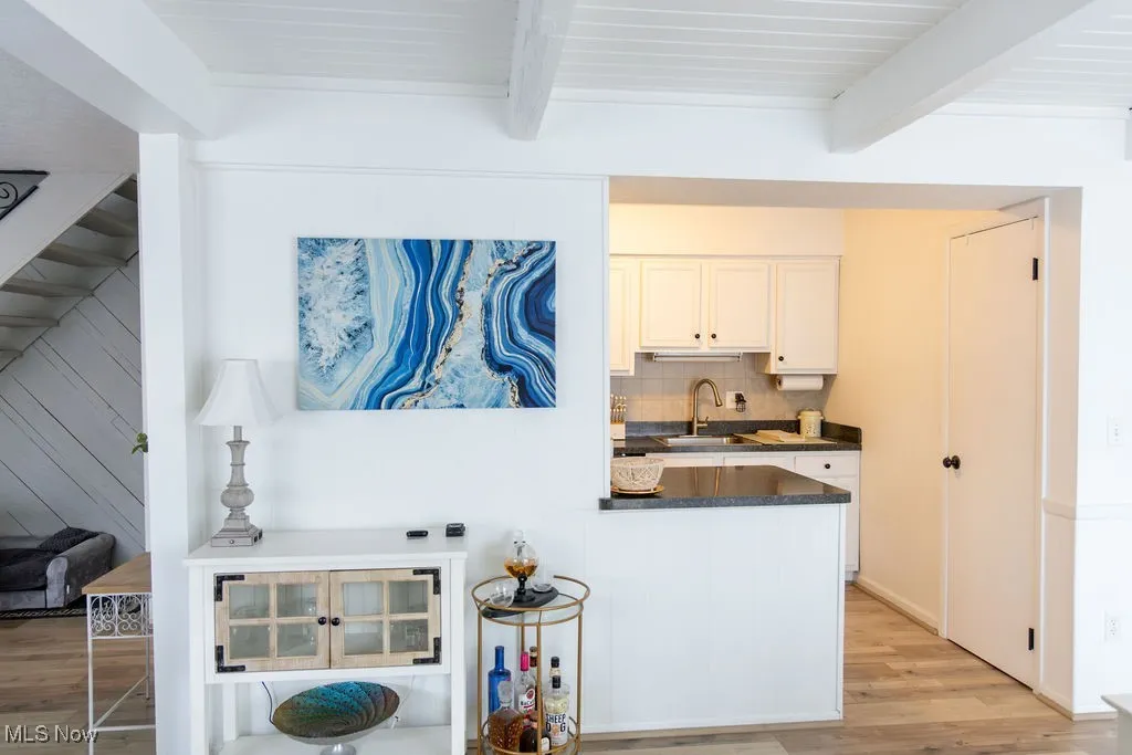 Kitchen featuring dark countertops, light wood-type flooring, white cabinets, beamed ceiling, and backsplash