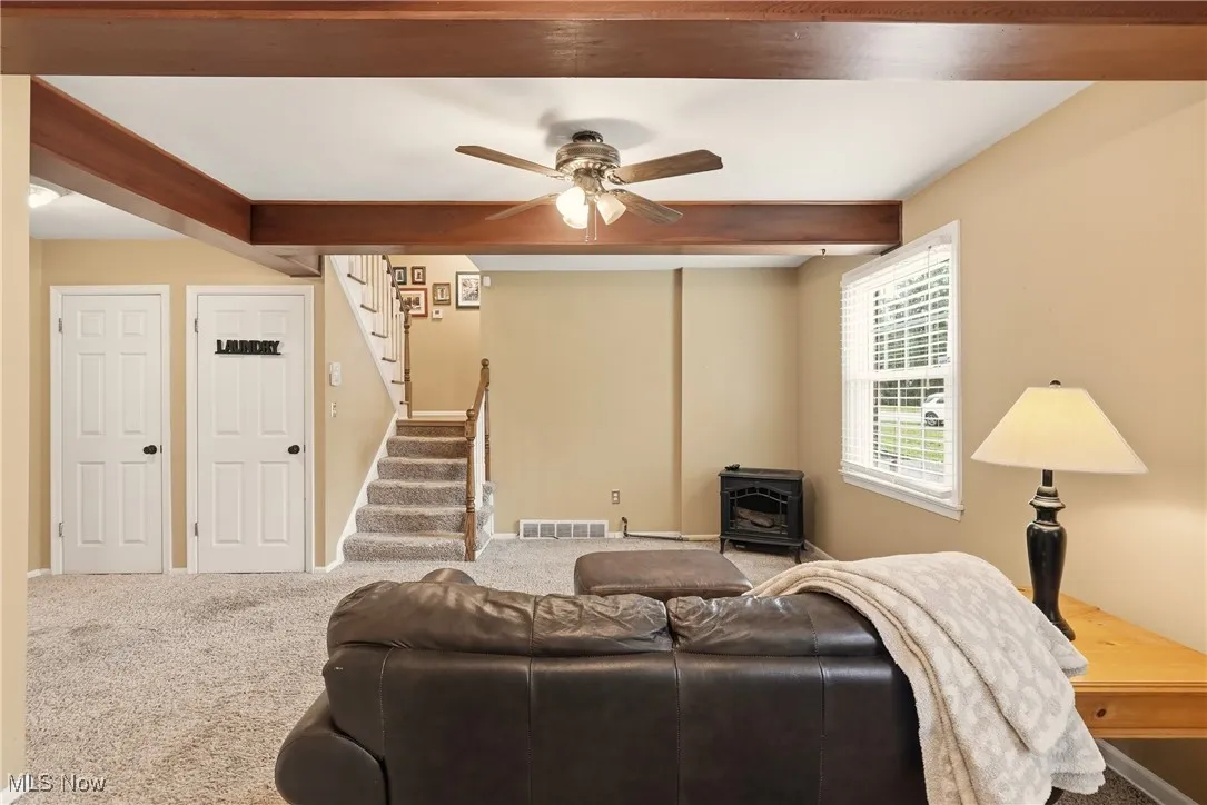 Living area with beam ceiling, carpet flooring, a wood stove, stairway, and a ceiling fan