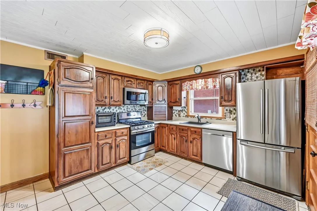 Kitchen featuring stainless steel appliances, brown cabinets, light countertops, ornamental molding, and light tile patterned floors
