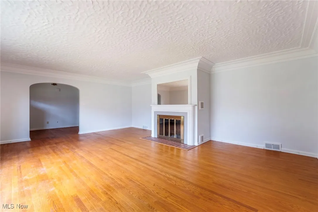 Unfurnished living room featuring arched walkways, a fireplace with flush hearth, ornamental molding, light wood-style flooring, and a textured ceiling