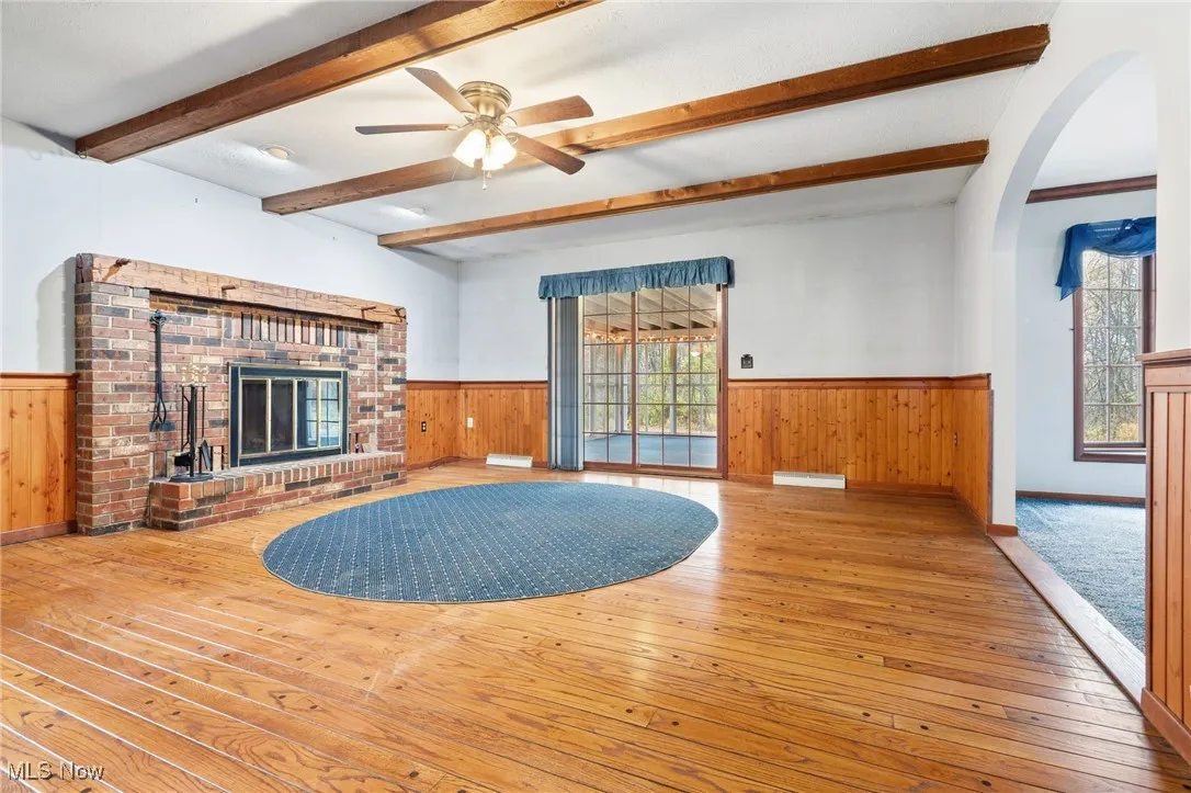 Unfurnished family room featuring wood-type flooring, a wainscoted wall, a ceiling fan, and a brick fireplace