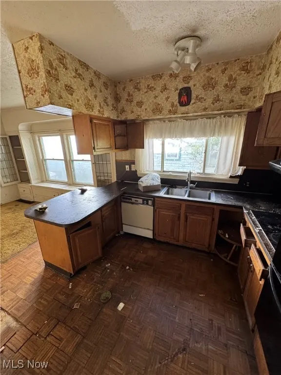 Kitchen with a textured ceiling, wallpapered walls, a peninsula, brown cabinets, and dishwasher