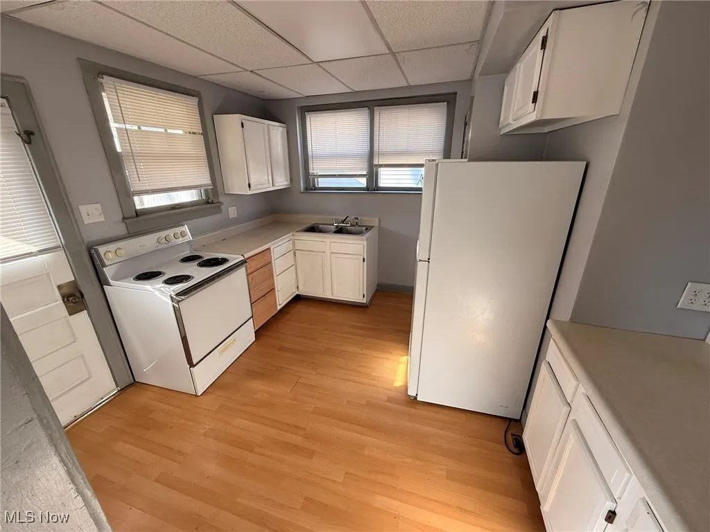 Kitchen with white appliances, white cabinets, a drop ceiling, light countertops, and light wood-type flooring
