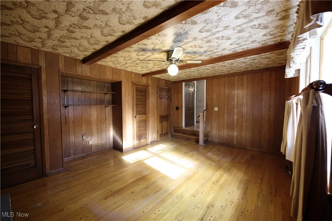 Unfurnished bedroom with beam ceiling, wooden walls, light wood finished floors, a ceiling fan, and two closets