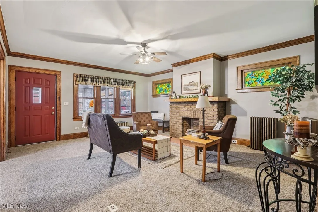 Living room featuring radiator heating unit, carpet, crown molding, a brick fireplace, and a ceiling fan