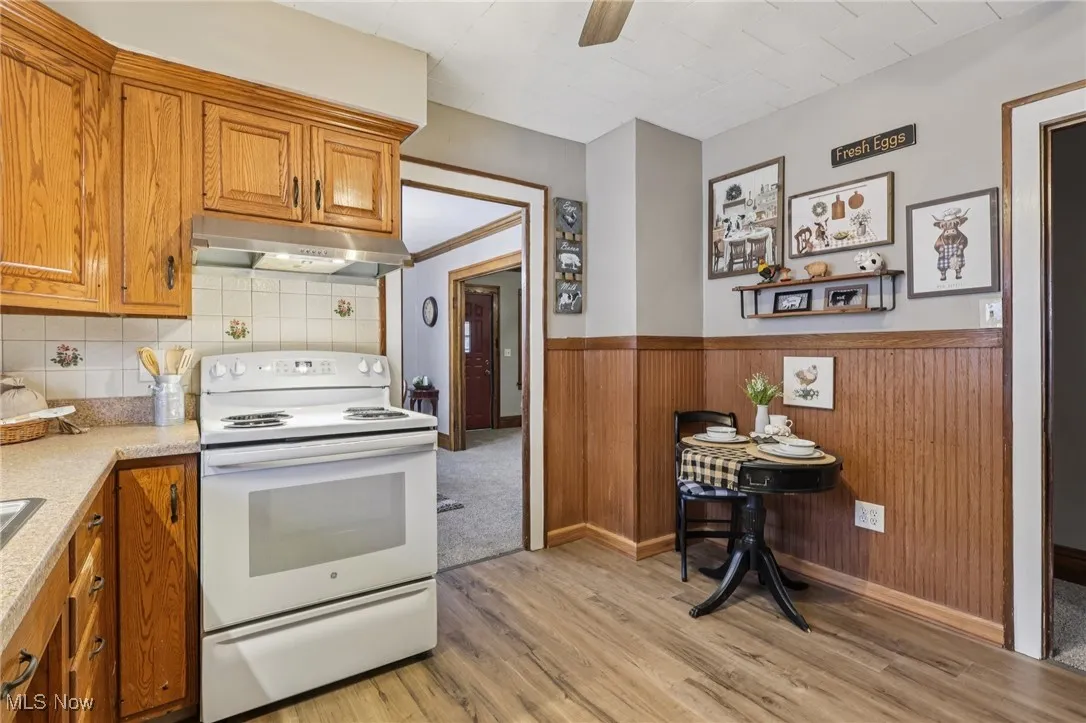 Kitchen with electric stove, brown cabinetry, wood walls, under cabinet range hood, and a wainscoted wall