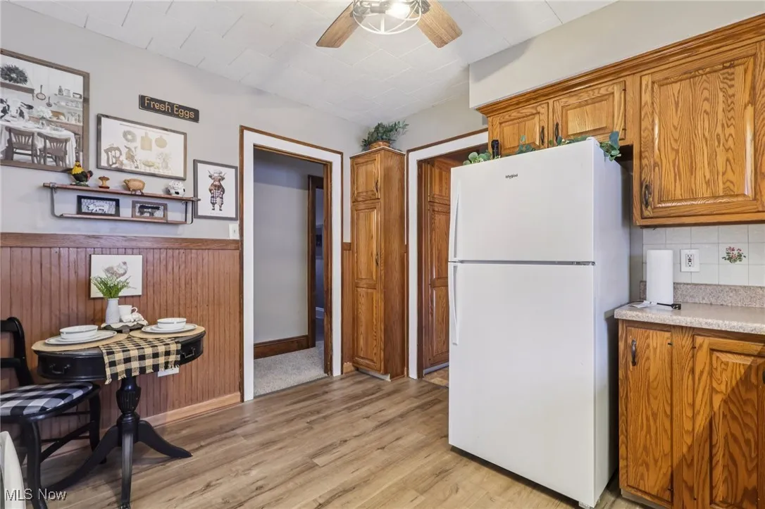 Kitchen featuring brown cabinetry, freestanding refrigerator, light countertops, ceiling fan, and a wainscoted wall