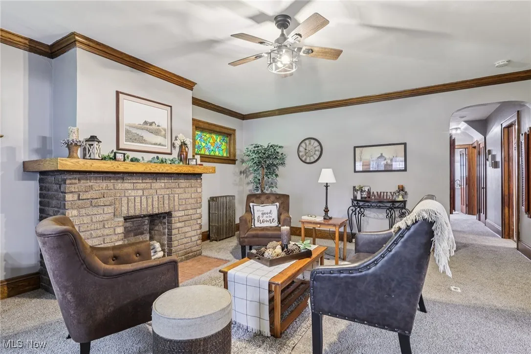 Carpeted living room featuring arched walkways, crown molding, a fireplace, a ceiling fan, and radiator heating unit
