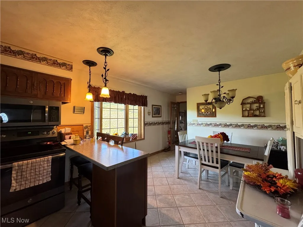 Kitchen featuring black range with electric stovetop, stainless steel microwave, a kitchen breakfast bar, decorative light fixtures, and light tile patterned flooring