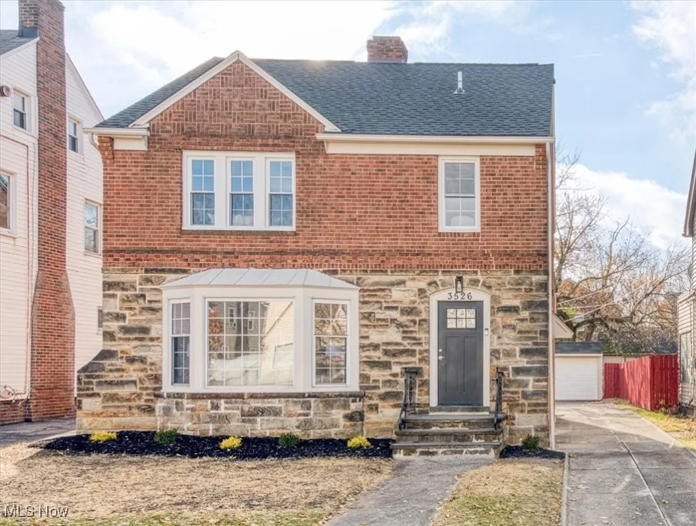 View of front facade with stone siding, a chimney, roof with shingles, and an outbuilding