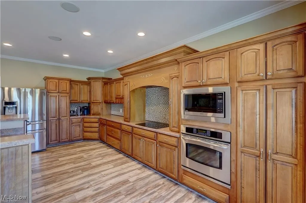 Kitchen with appliances with stainless steel finishes, ornamental molding, backsplash, light wood finished floors, and brown cabinets