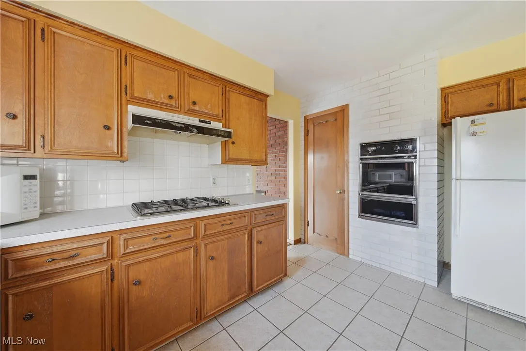 Kitchen with brown cabinetry, light countertops, white appliances, tasteful backsplash, and under cabinet range hood