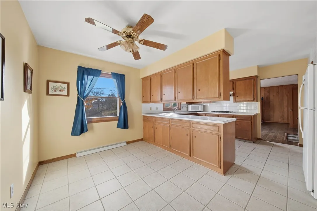 Kitchen featuring a baseboard radiator, a peninsula, white appliances, a ceiling fan, and decorative backsplash