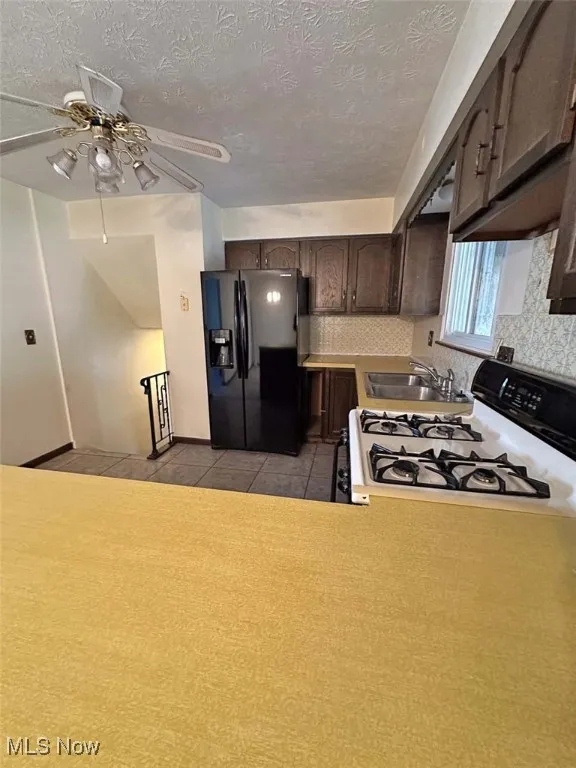 Kitchen with black refrigerator with ice dispenser, white gas range oven, dark brown cabinetry, light countertops, and a textured ceiling