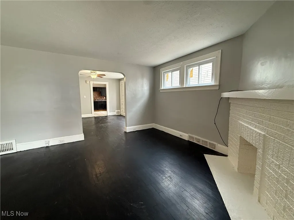 Unfurnished living room featuring a brick fireplace, arched walkways, a textured ceiling, dark wood finished floors, and ceiling fan