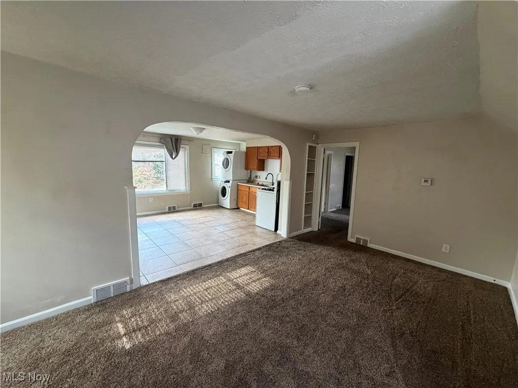 Unfurnished living room featuring arched walkways, a textured ceiling, light colored carpet, light tile patterned floors, and stacked washer and clothes dryer