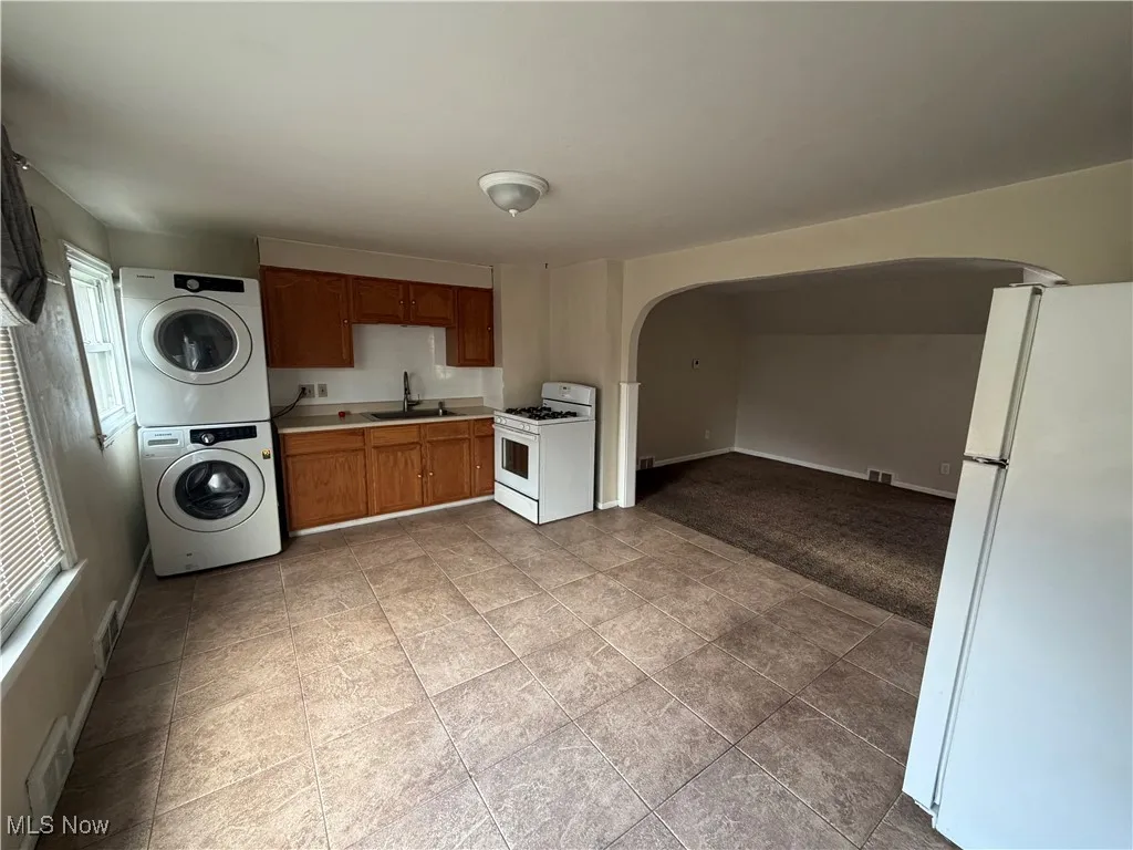 Kitchen with white appliances, light countertops, arched walkways, stacked washer / dryer, and brown cabinets
