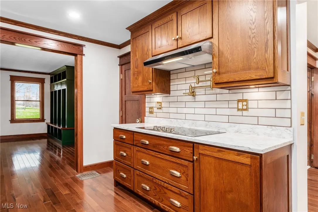 Kitchen with ornamental molding, brown cabinets, dark wood-style flooring, under cabinet range hood, and light stone counters