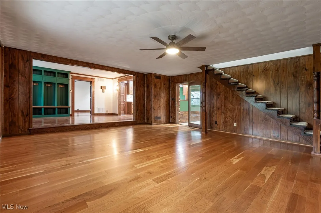 Unfurnished living room featuring wood walls, light wood-style flooring, stairs, and ceiling fan