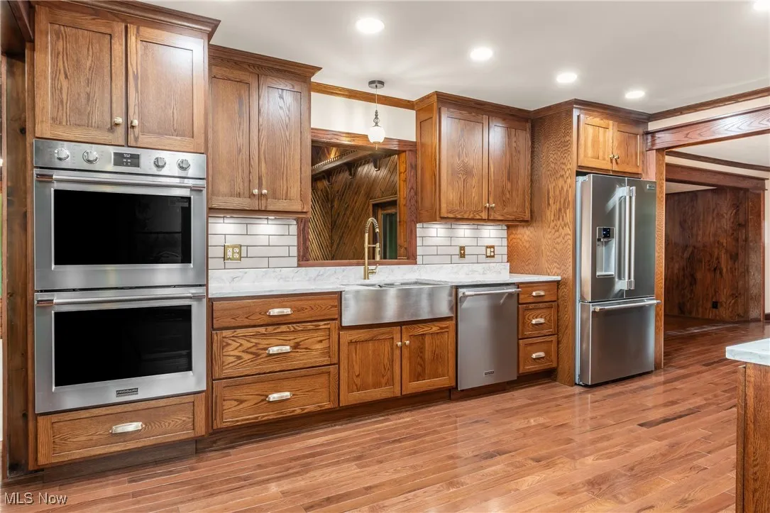 Kitchen with stainless steel appliances, brown cabinetry, decorative backsplash, light wood-style flooring, and light stone countertops