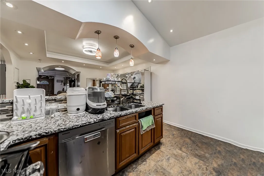 Kitchen featuring light stone countertops, dishwasher, pendant lighting, a raised ceiling, and brown cabinetry