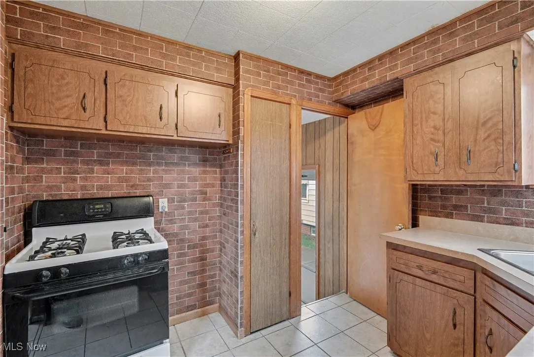 Kitchen featuring black gas range oven, light countertops, light tile patterned floors, brown cabinets, and brick wall