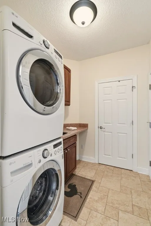 Washroom with stacked washing machine and dryer, a textured ceiling, cabinet space, and light stone finish flooring