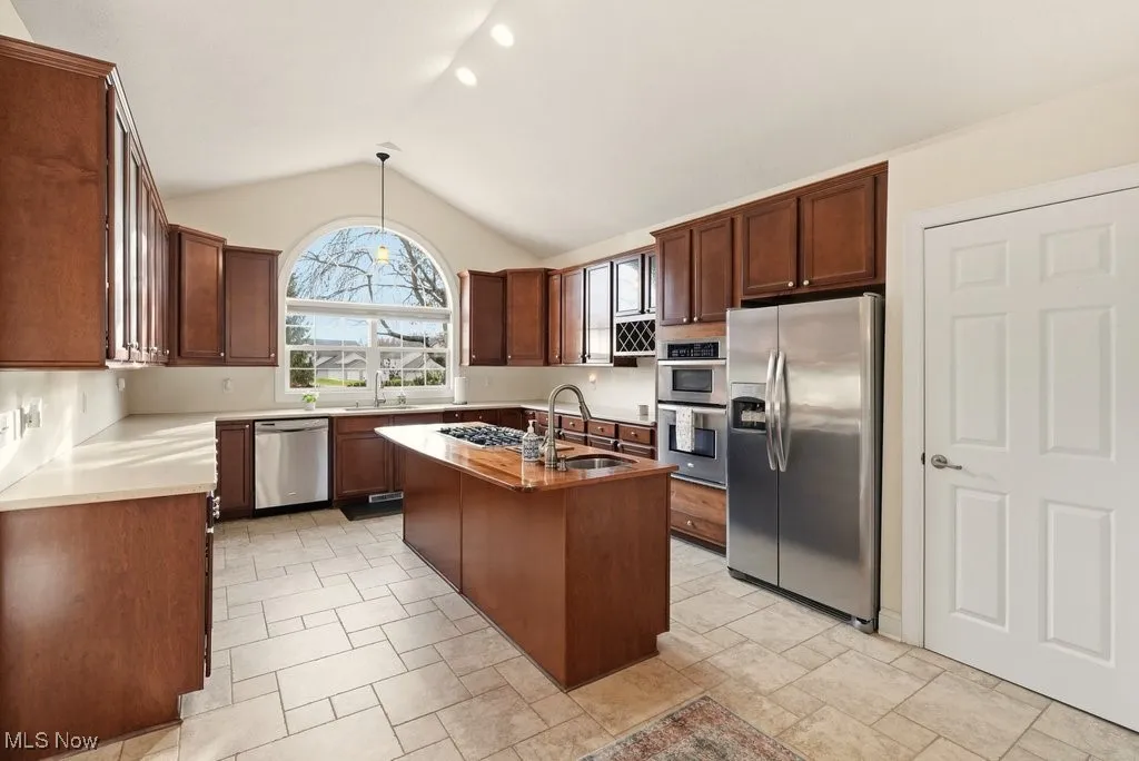 Kitchen featuring stainless steel appliances, decorative light fixtures, vaulted ceiling, a center island with sink, and stone tile floors
