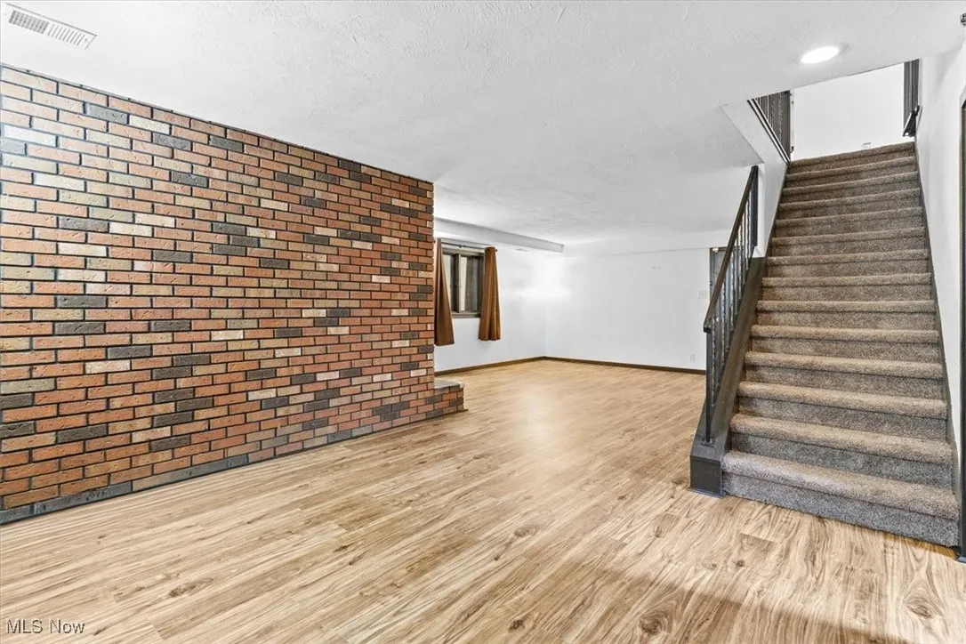 Unfurnished living room featuring brick wall, light wood-type flooring, stairs, and a textured ceiling