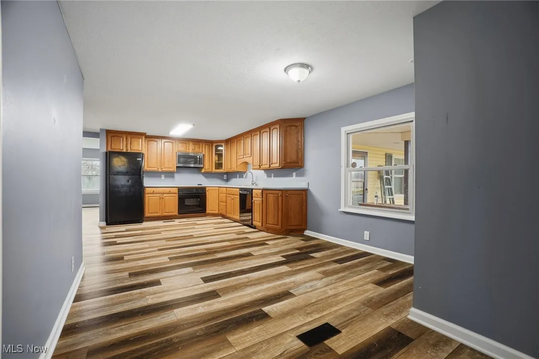 Kitchen with glass insert cabinets, black appliances, brown cabinetry, light countertops, and dark wood-type flooring