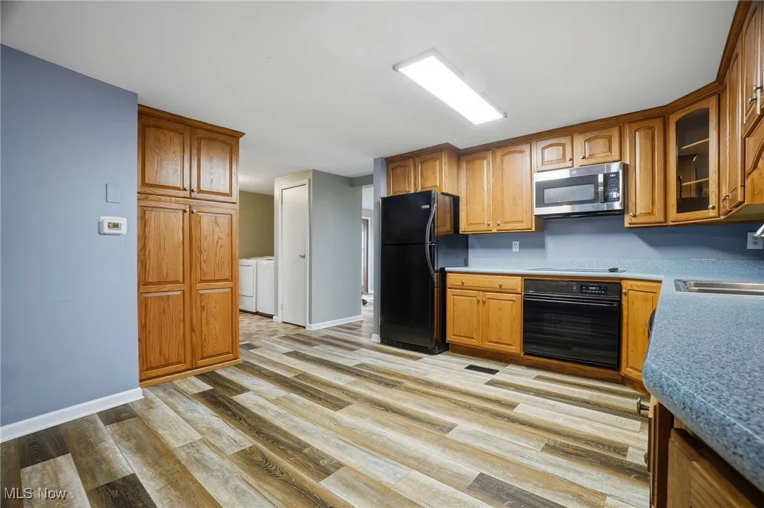 Kitchen with black appliances, glass insert cabinets, light wood finished floors, brown cabinetry, and washer and clothes dryer