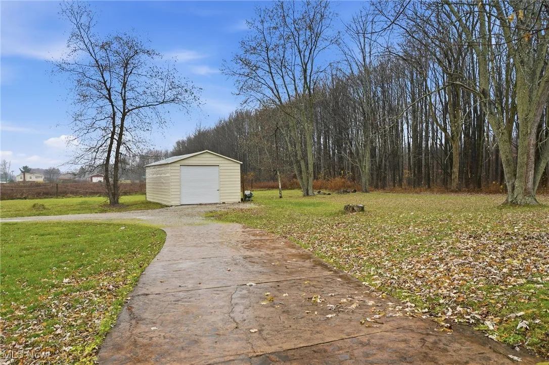 View of grassy yard with an outdoor structure, driveway, a detached garage, and a view of trees