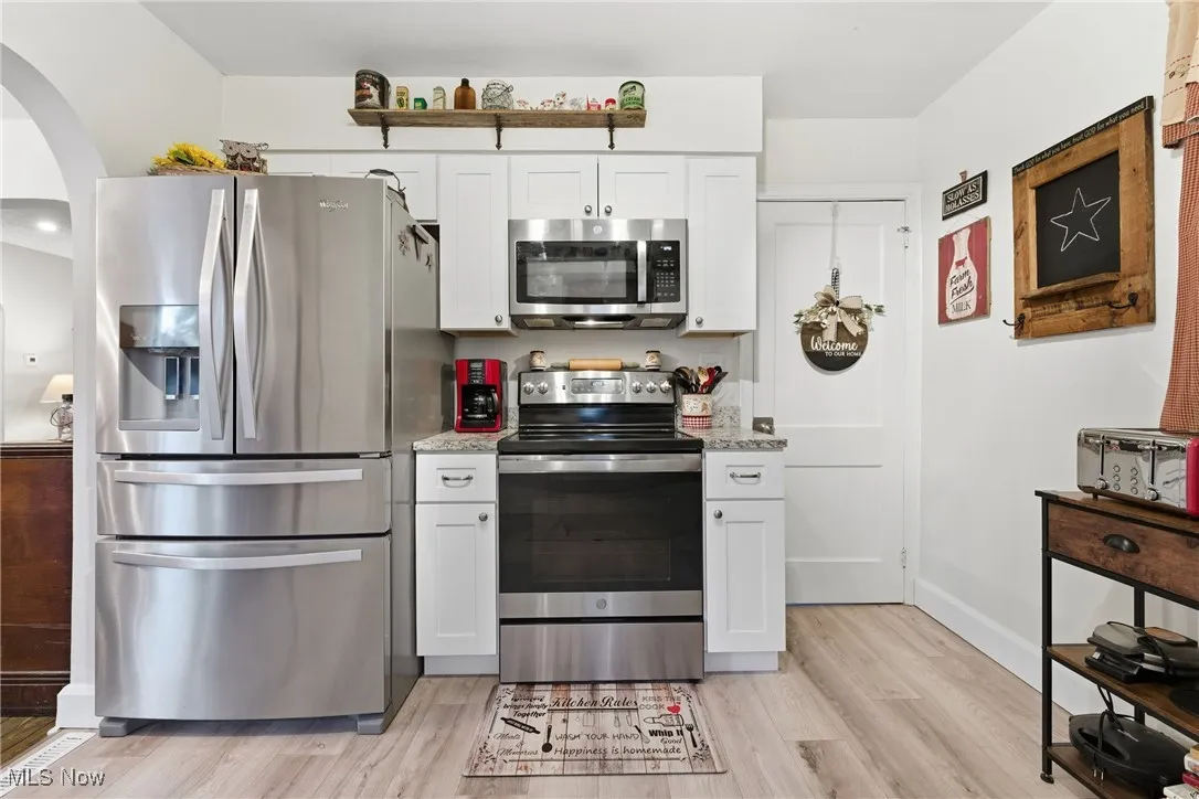 Kitchen with appliances with stainless steel finishes, white cabinetry, light wood-style floors, arched walkways, and light stone counters