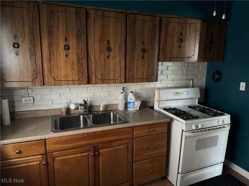 Kitchen featuring white gas stove, tasteful backsplash, brown cabinetry, and dark countertops