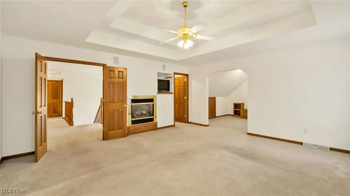  Master bedroom featuring light colored carpet, a ceiling fan, a tray ceiling, and a gas fireplace