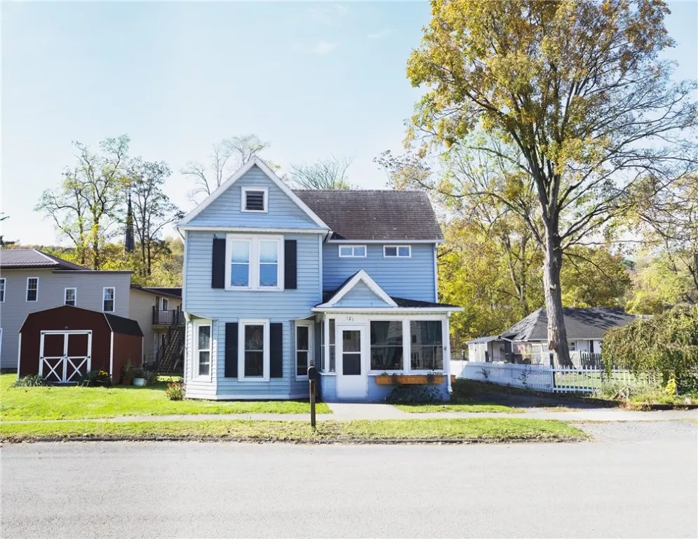 Classic two-story village home with light blue siding, black shutters, and a welcoming enclosed front porch. Large Storge Shed is included.