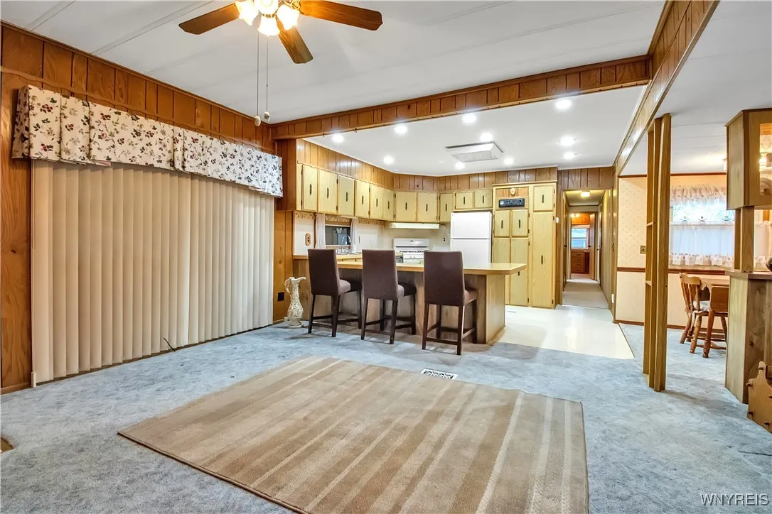 Carpeted Living room with windowed wall + ceiling fan overlooks Kitchen