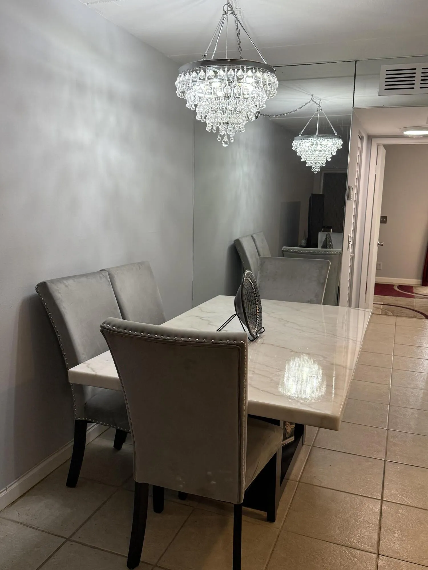 Elegant crystal chandelier illuminating a dining area featuring a rectangular marble-finish table, upholstered dining chairs with stud detailing, and tile flooring