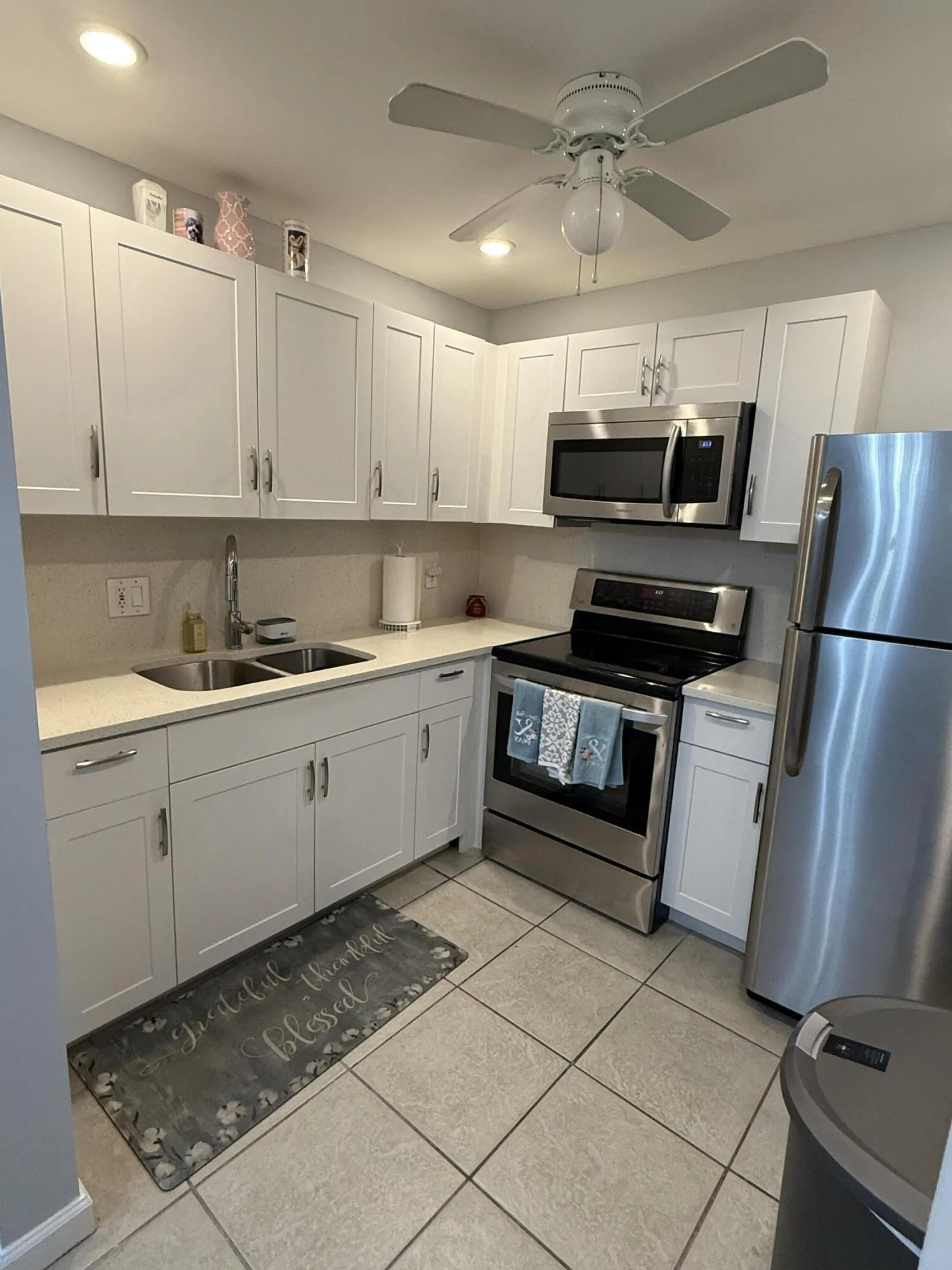 Modern kitchen featuring white shaker cabinetry, stainless steel appliances, light-toned countertops, a double basin sink, and ceiling fan with integrated lighting