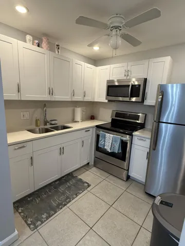 Modern kitchen featuring white shaker cabinetry, stainless steel appliances, light-toned countertops, a double basin sink, and ceiling fan with integrated lighting
